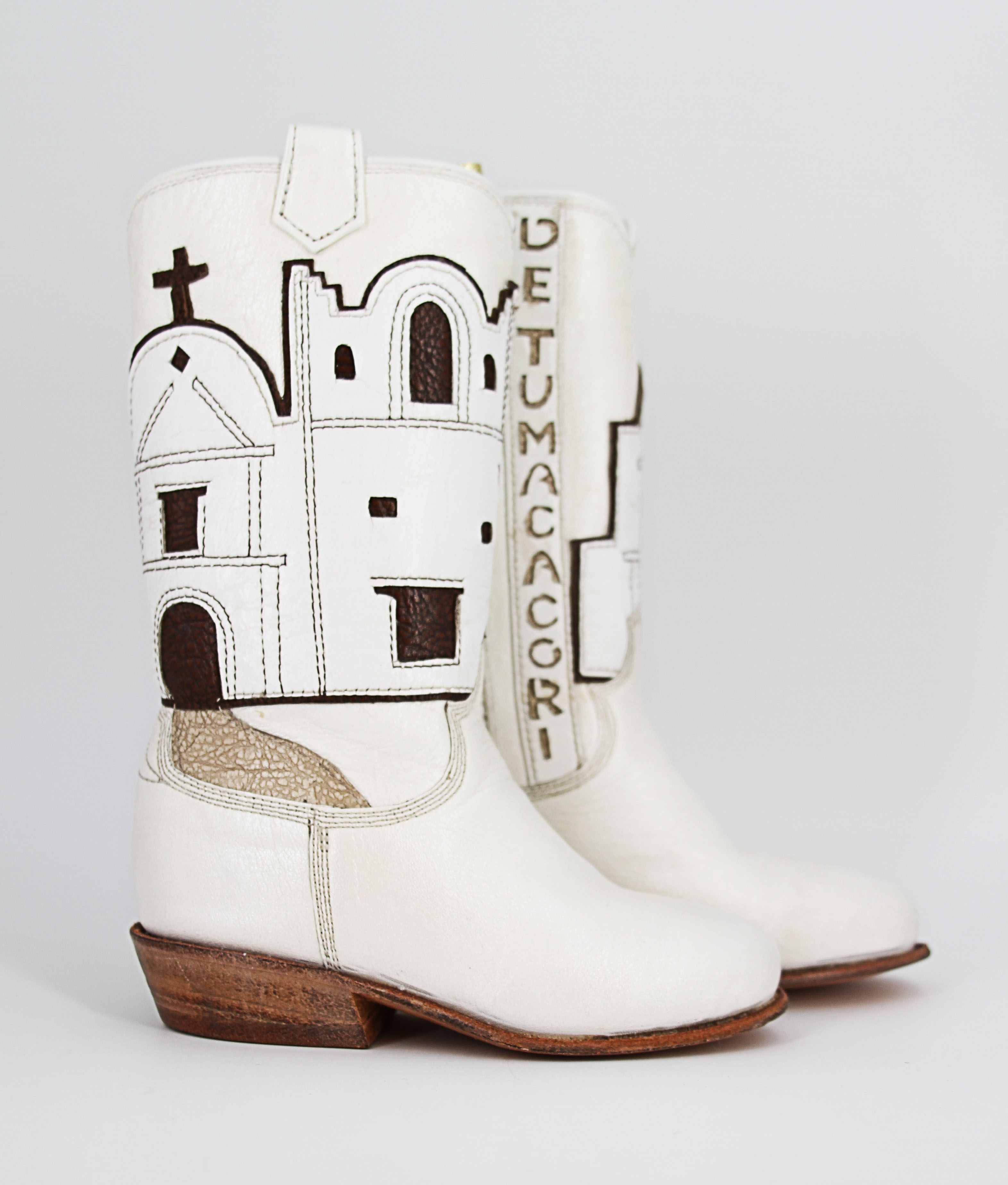 Miniature white leather boots with cutout name, "San Cayentano de Tumacacori," leatherwork depiction of the mission, and a gold leather cord for hanging. Signed inside by master craftsman Paul Bond, AZ, of the Paul Bond Boot Company.
